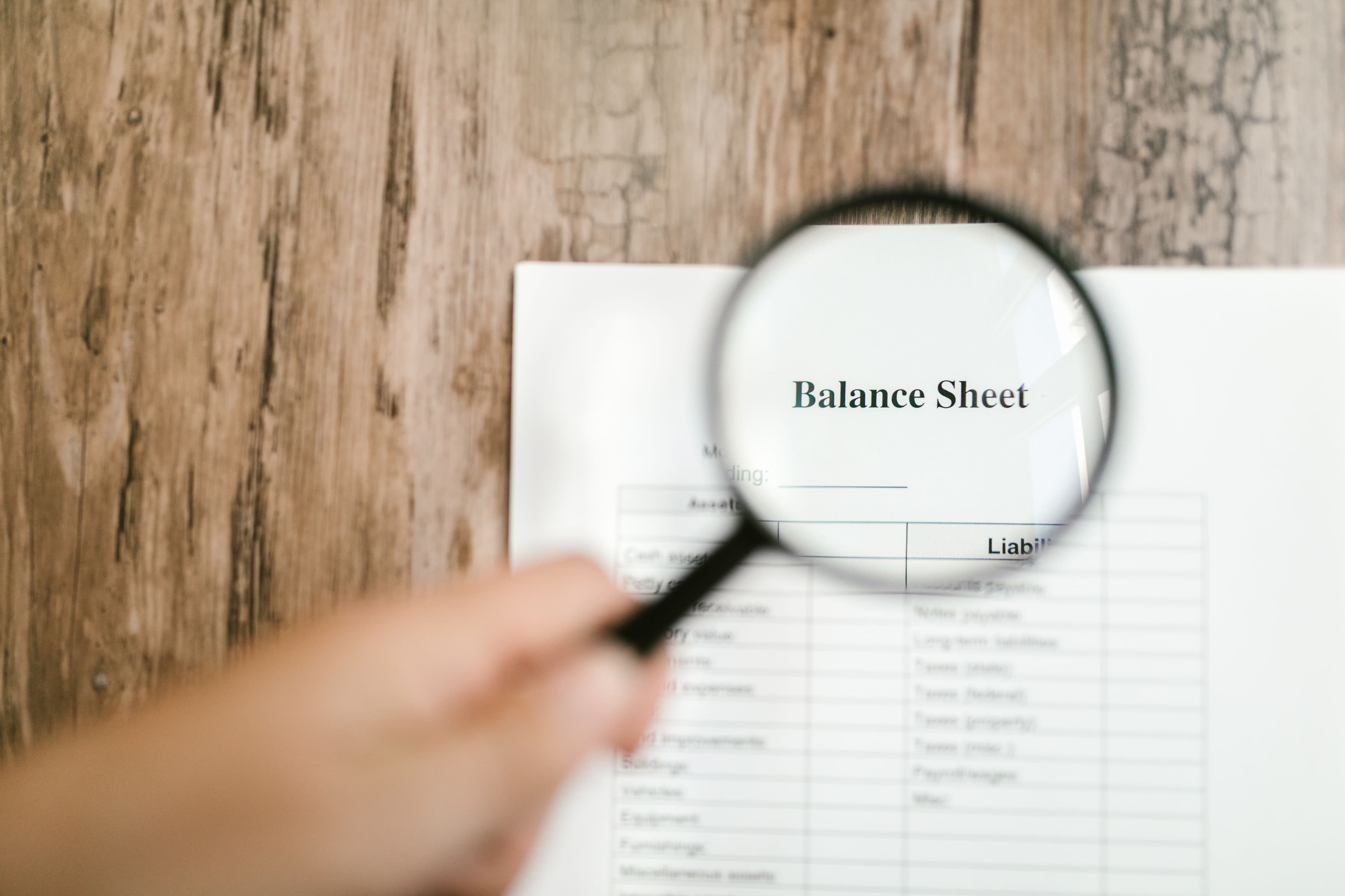 A hand holding a magnifying glass over a printed balance sheet on a wooden desk.