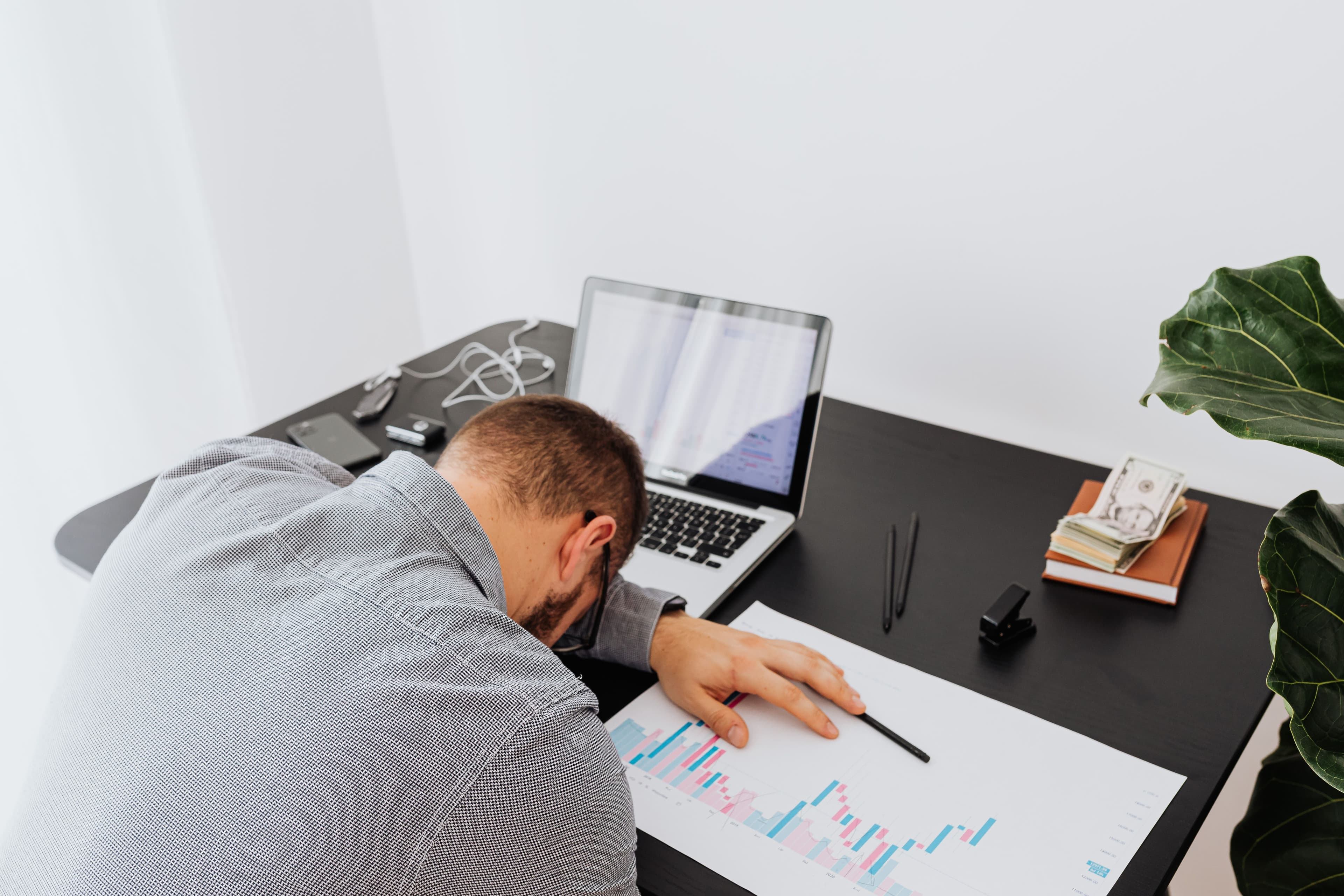 A man slumped head-down over his desk beside a laptop and a printed bar chart, worn out by paperwork.
