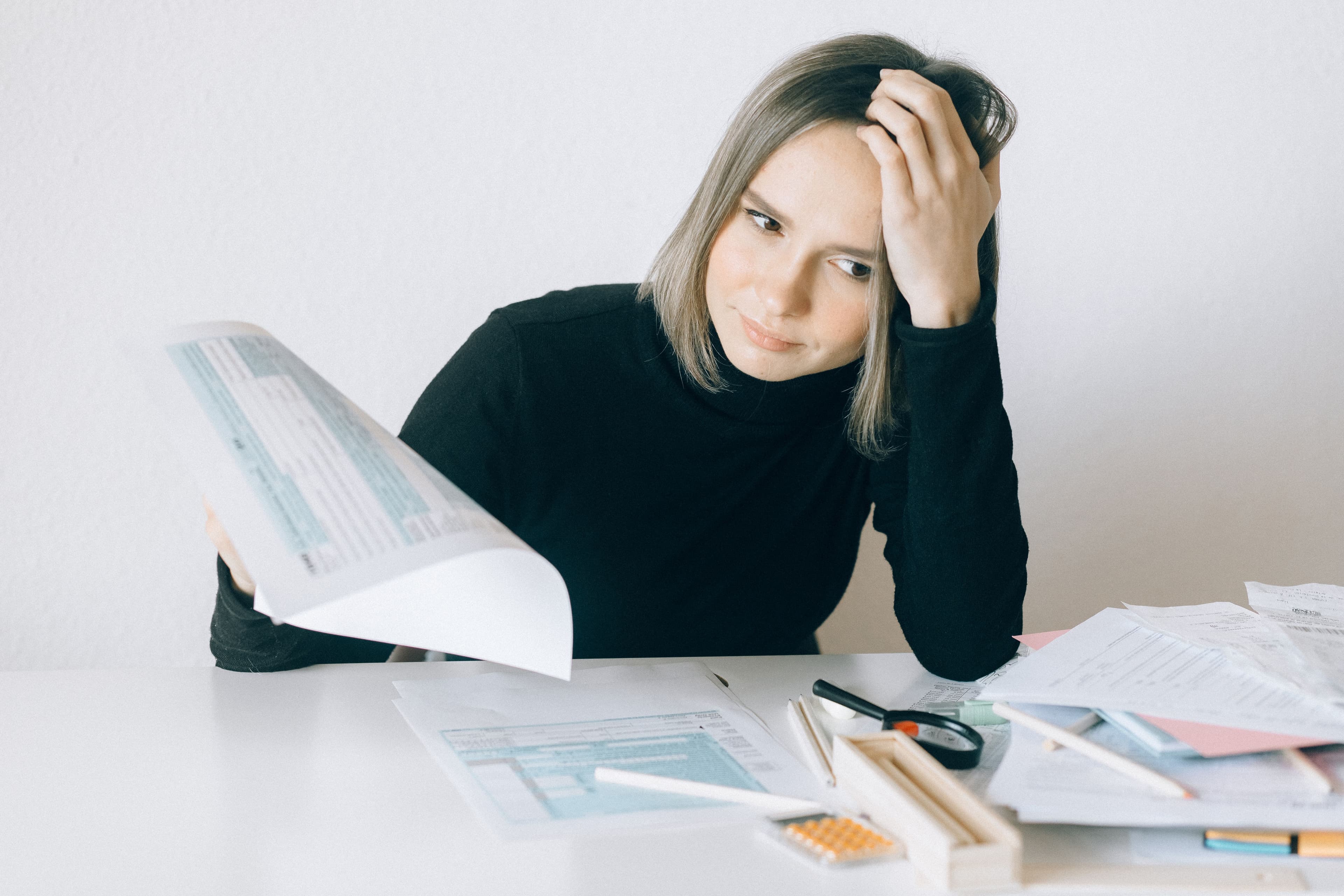 A woman resting her head on her hand while reading a long tax form, with more forms and a calculator spread across the desk.