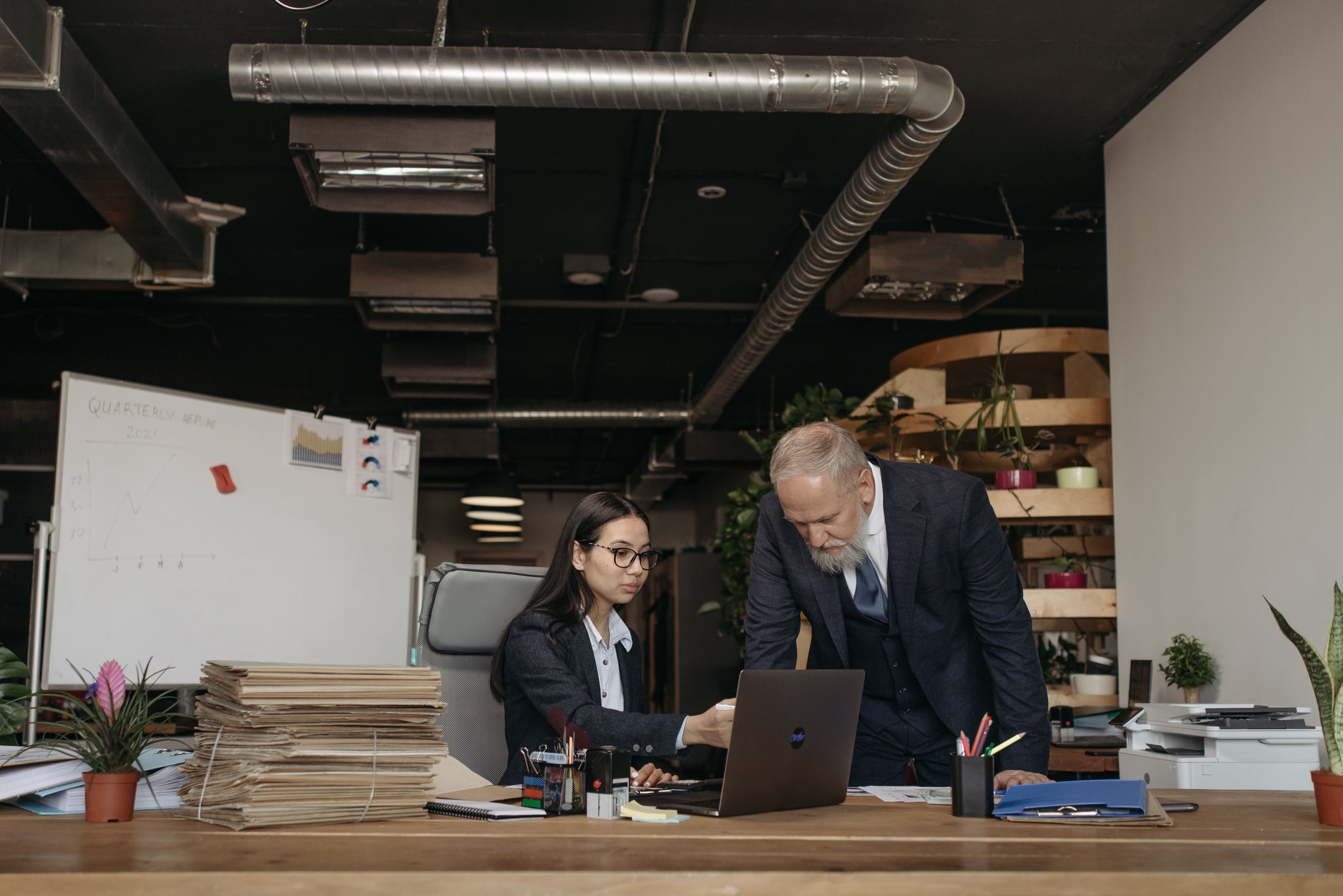 A senior manager leaning over a colleague's laptop in an open-plan office, with a quarterly report chart sketched on the whiteboard behind them.
