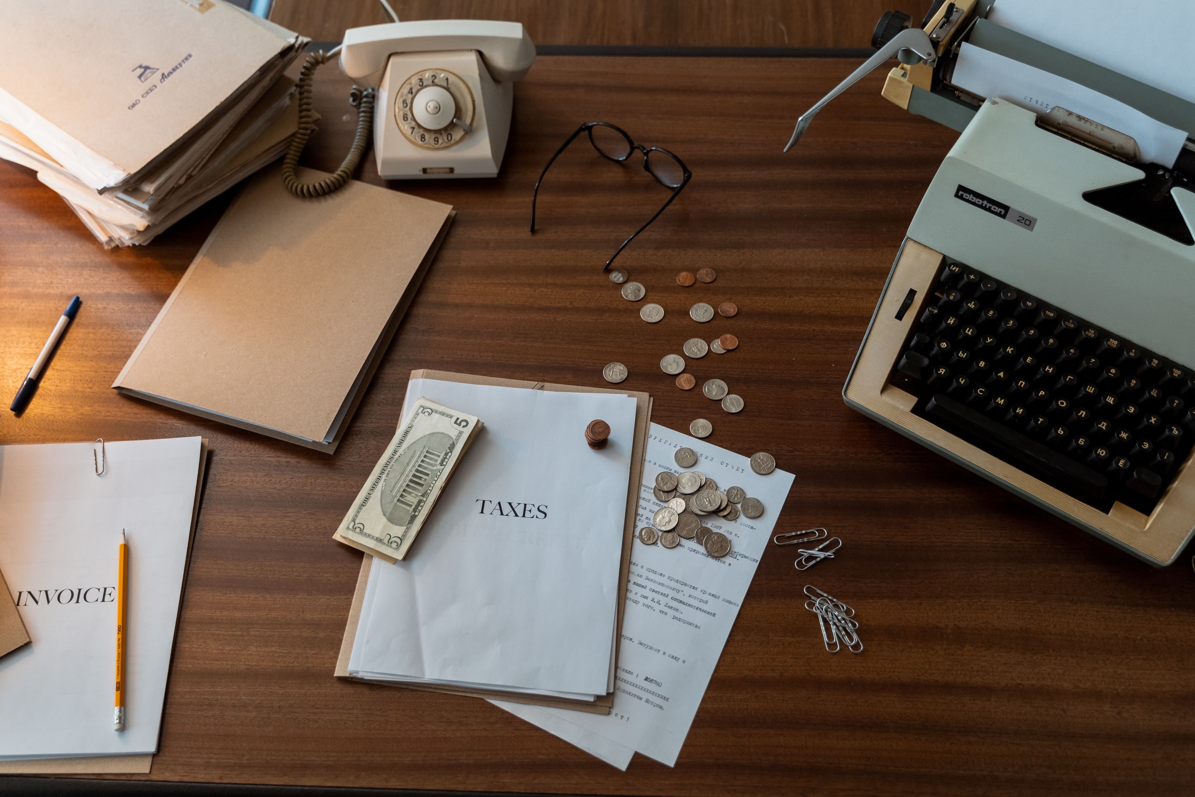An old-fashioned desk with a rotary telephone, a typewriter, paper invoices, a folder marked "Taxes" and loose coins.