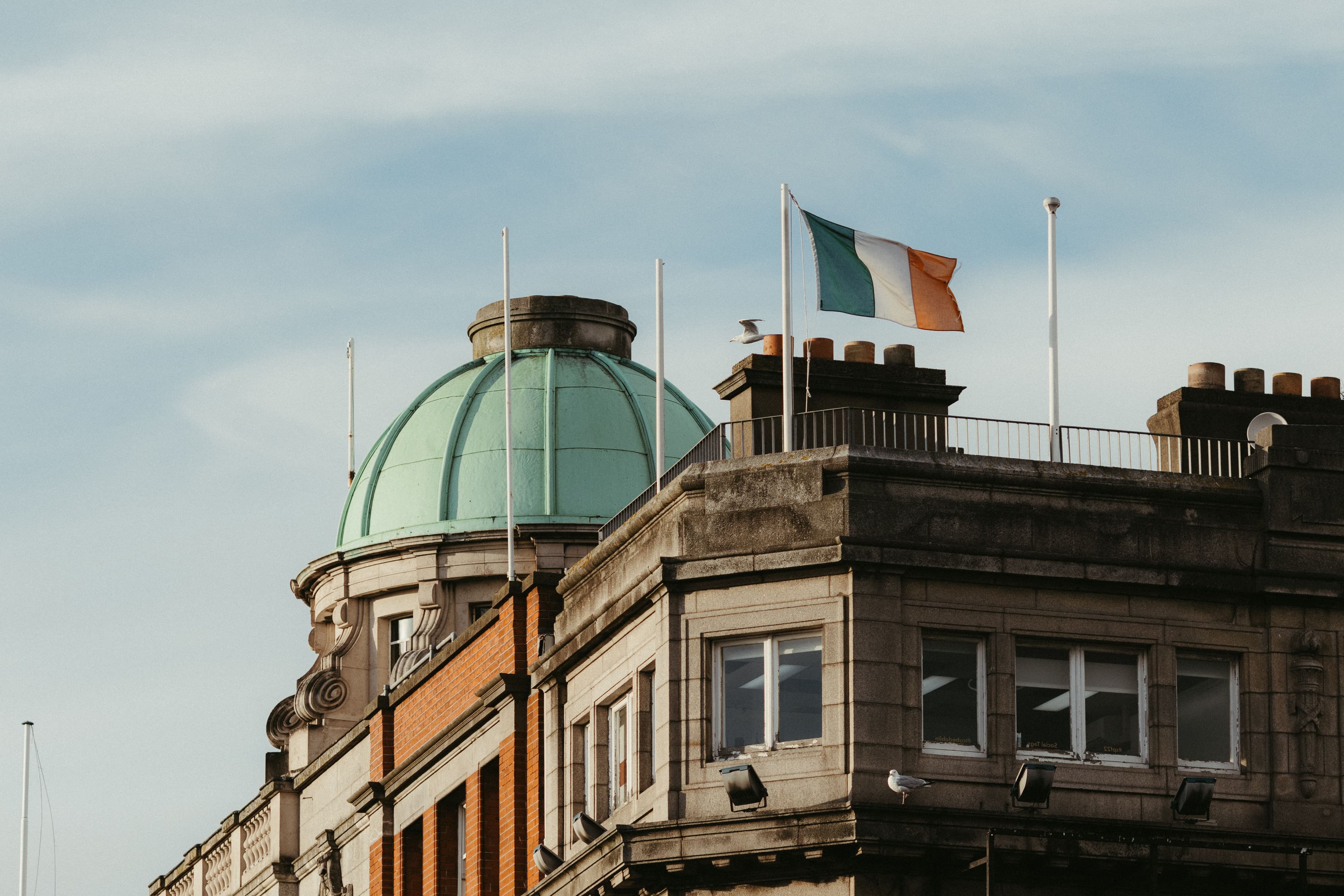 The Irish tricolour flag waving on the rooftop of a classic stone corporate building featuring a distinctive green copper dome under a clear sky.