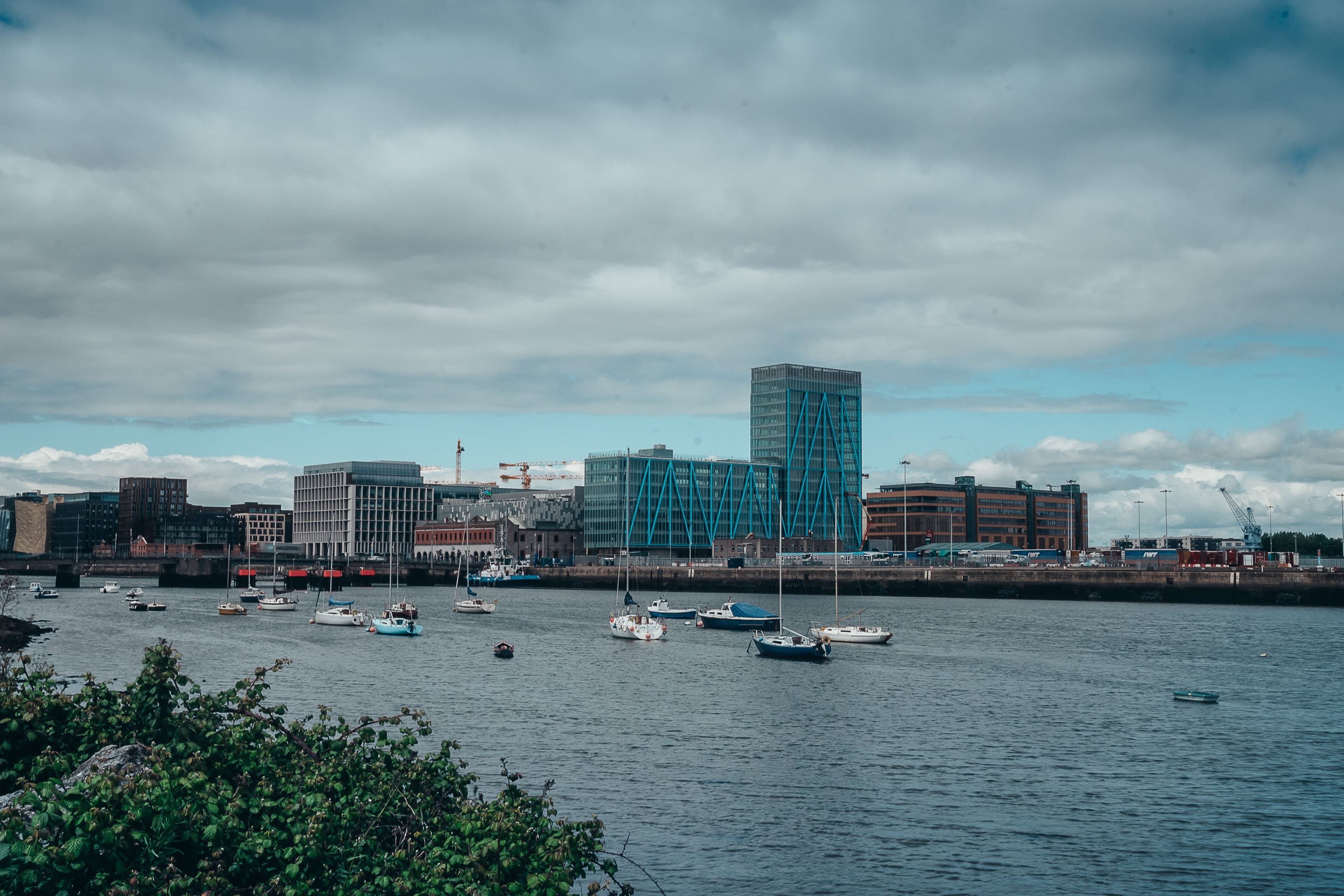 Sailing boats moored on the water in front of modern glass office blocks and construction cranes in Dublin's docklands.