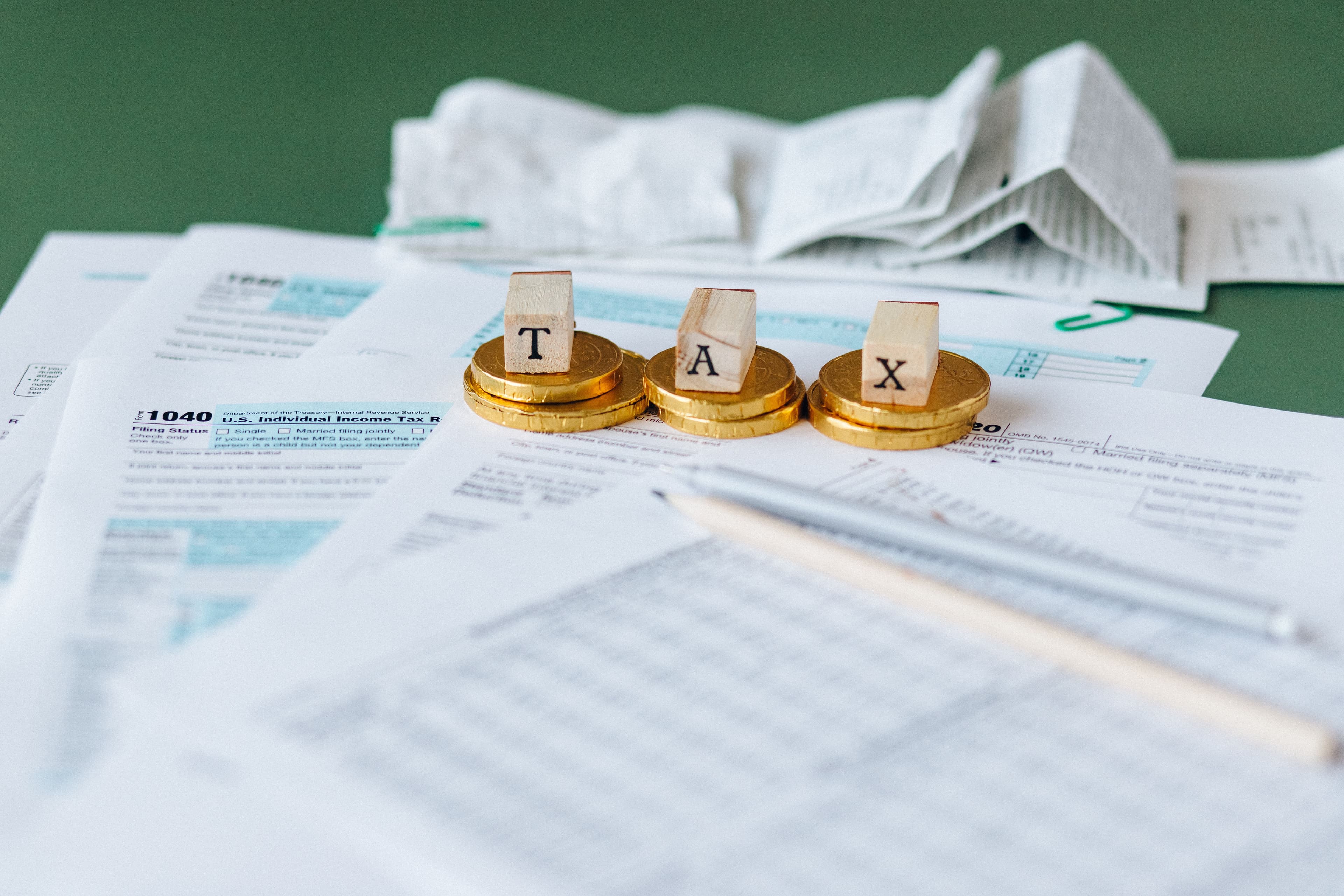 Wooden letter blocks spelling "TAX" standing on stacks of gold coins on top of tax return forms.